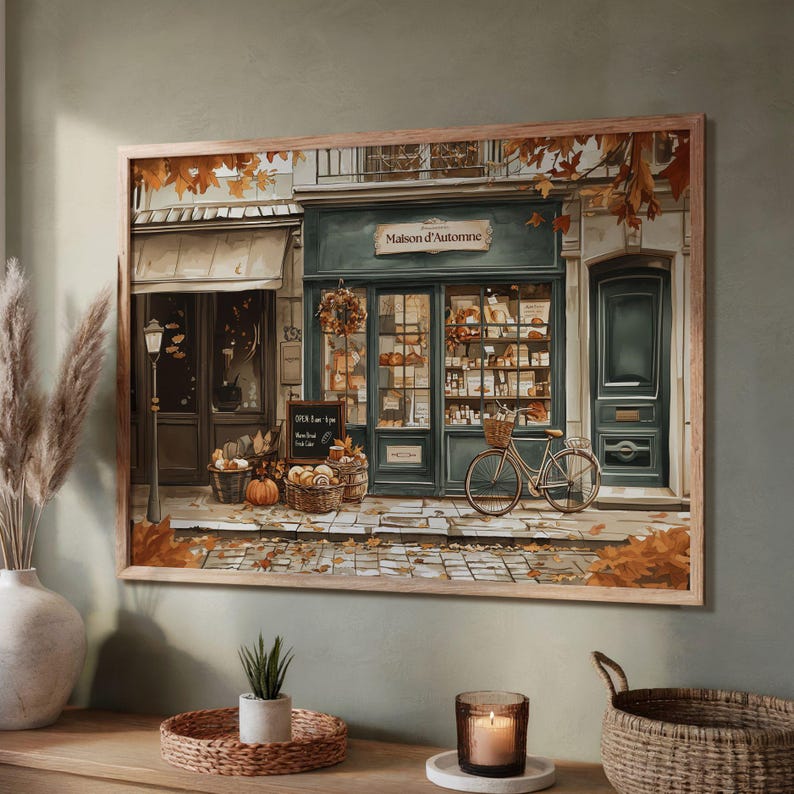 May include: A framed print of a quaint bakery, "Maison d'Automne", with a teal facade and a bicycle parked outside. Baskets of pumpkins and bread sit on the sidewalk, surrounded by autumn leaves. The artwork evokes a cozy, autumnal atmosphere.