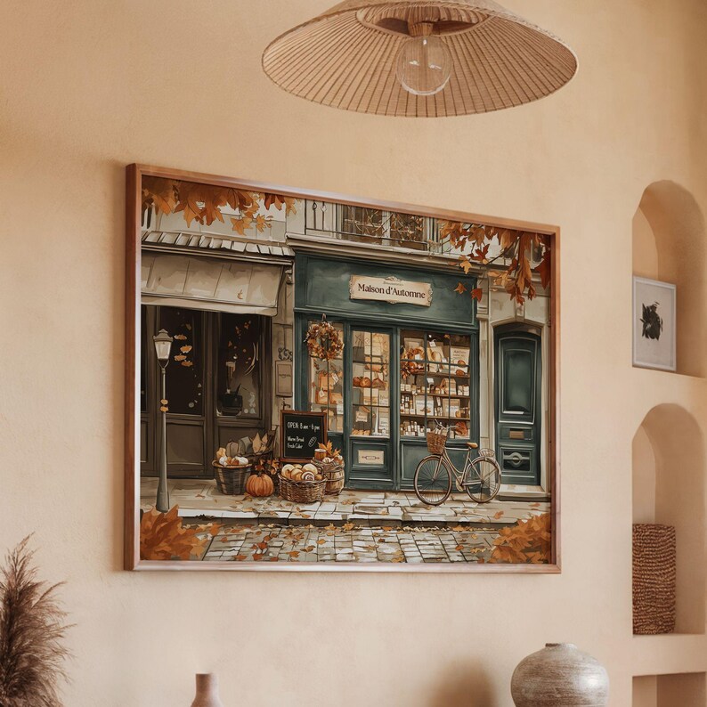 May include: A framed print of a shop with the text "Maison d'Automne" above the door. The shop has a bicycle parked outside and baskets of pumpkins and bread. The artwork is in warm autumn colors.