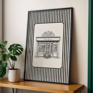 May include: Black and white framed print featuring a detailed illustration of a patisserie shop. The artwork includes the word "PATISSERIE" above the shop's awning and "Cakes & Macarons" below. The frame has a black and white striped border.