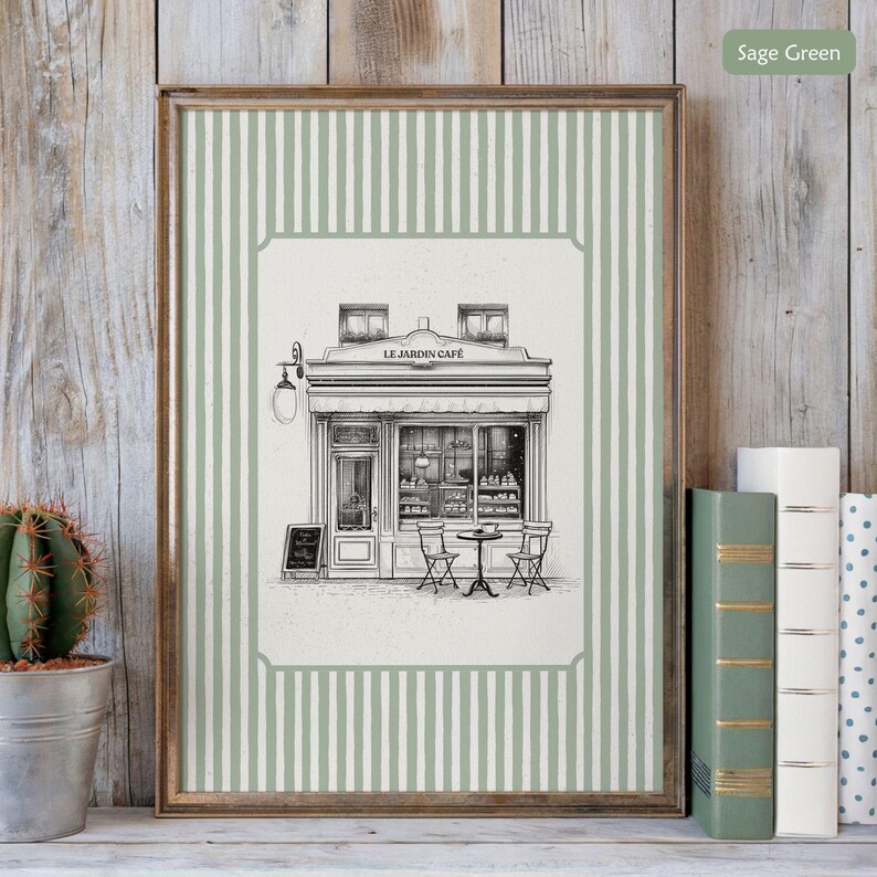 May include: Framed art print featuring a detailed black and white illustration of a cafe, "Le Jardin Cafe", with outdoor seating. The frame has a sage green and white striped border. A small cactus in a metal pot is on the left.