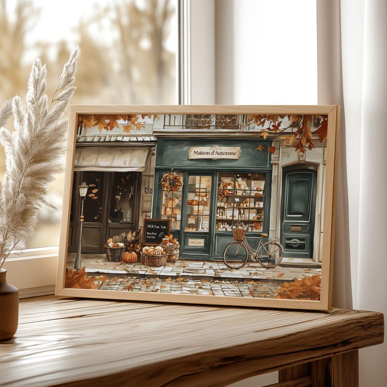 May include: A framed print of a quaint French bakery, "Maison d'Automne," with a teal facade, autumn leaves, and a bicycle. The image features baskets of produce, pumpkins, and a sign indicating opening hours. The artwork is set on a wooden table.