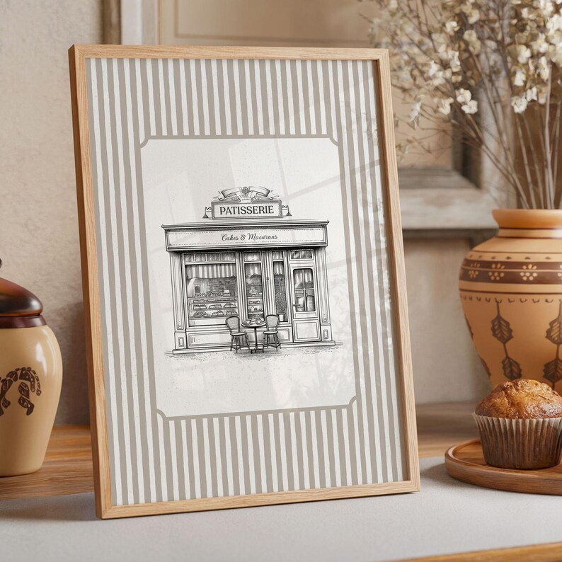 May include: A framed print featuring a black and white illustration of a French patisserie with the text "Patisserie Cakes & Macarons." The frame has a light wood finish, and the background has vertical gray and white stripes.