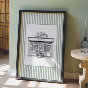 May include: Black framed print featuring a detailed black and white illustration of a French patisserie. The print includes the word "PATISSERIE" above the shop and "Cakes & Macarons" below. The background has a green and white striped pattern.