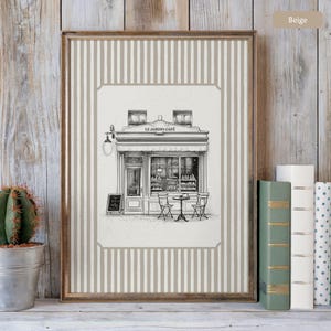 May include: Framed print featuring a detailed black and white illustration of a cafe, "Le Jardin Cafe", with outdoor seating. The print has a beige striped background and a wooden frame. A small cactus in a metal pot is on the left.