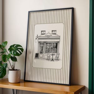 May include: Framed print featuring a detailed black and white illustration of a cafe facade with the text "LE JARDIN CAFE". The print is set against a beige and white striped background, framed in black. A potted plant sits on a wooden surface.