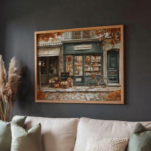 May include: A framed print of a quaint French shop, "Maison d'Automne", with a teal facade, a bicycle, and autumn decor. The artwork features a cobblestone street, pumpkins, and fall foliage, creating a warm, inviting scene. The frame is made of light brown wood.