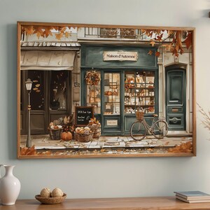 May include: A framed print of a quaint bakery, "Maison d'Automne," with a teal facade, a bicycle, and baskets of bread. Autumn leaves and pumpkins add to the cozy, seasonal atmosphere. A sign indicates the bakery's hours.