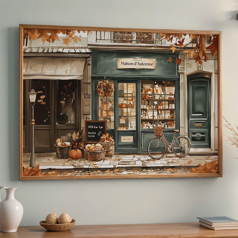 May include: A framed print of a quaint bakery, "Maison d'Automne," with a teal facade, a bicycle, and baskets of bread. Autumn leaves and pumpkins add to the cozy, seasonal atmosphere. A sign indicates the bakery's hours.