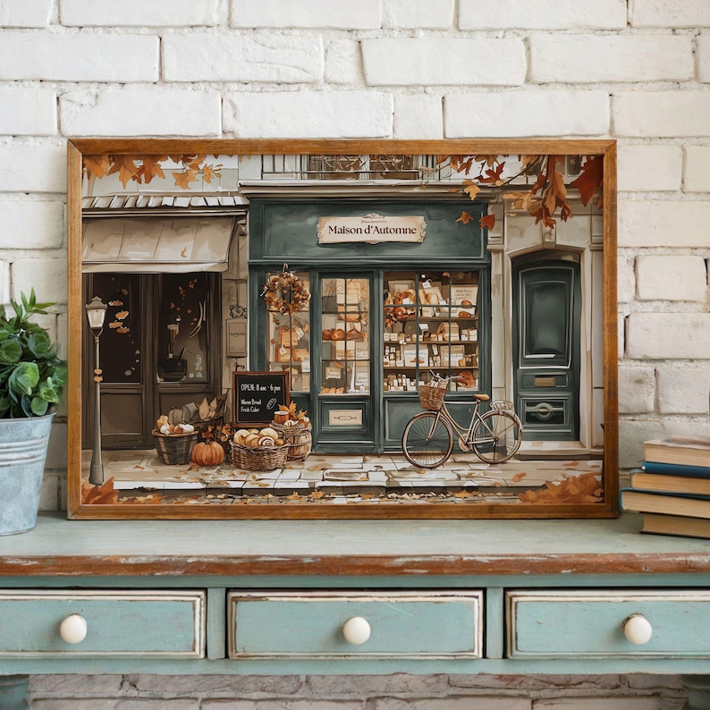 May include: A framed print depicts a quaint bakery, "Maison d'Automne," with a teal facade and autumn decor. Baskets of pumpkins and bread sit outside, with a bicycle parked nearby. The image is set against a brick wall.