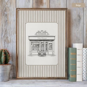 May include: Framed print featuring a black and white illustration of a patisserie shop with the text "PATISSERIE Cakes & Macarons." The print has a beige and white striped border and a wooden frame. A small cactus in a metal pot and books are visible.