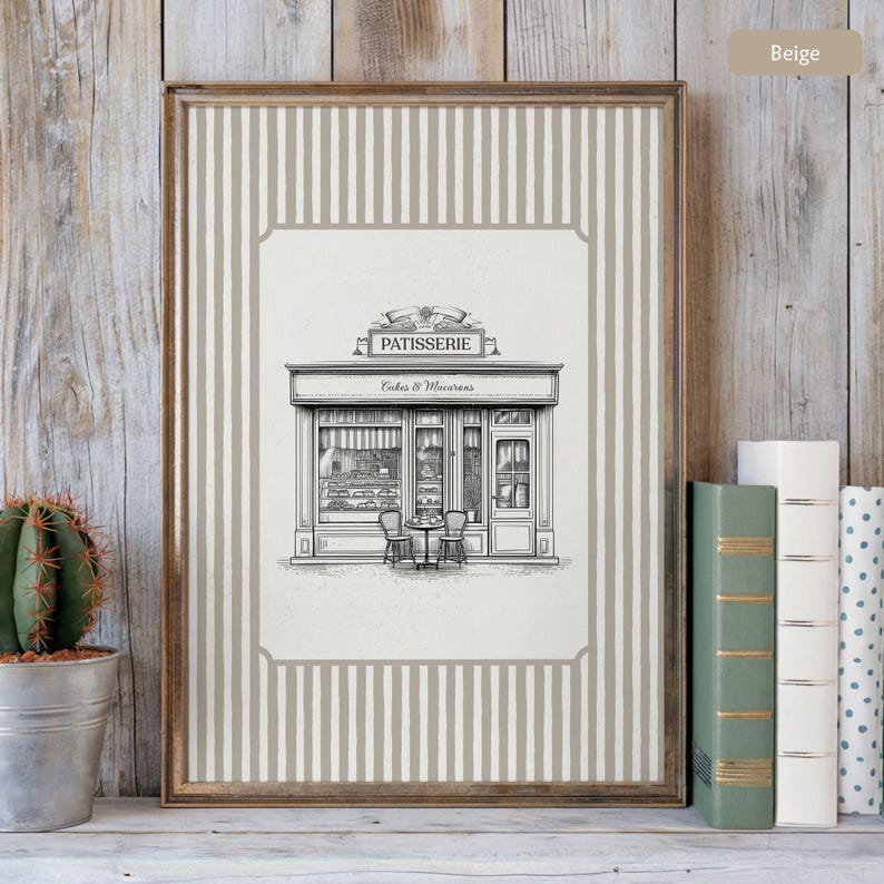 May include: Framed print featuring a black and white illustration of a patisserie shop with the text "PATISSERIE Cakes & Macarons." The print has a beige and white striped border and a wooden frame. A small cactus in a metal pot and books are visible.