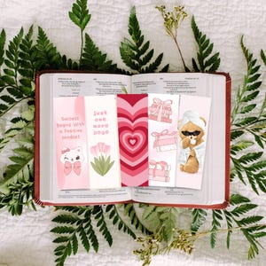 Könnte beinhalten: Vier rosa und weiße Lesezeichen mit verschiedenen Designs. Ein Lesezeichen zeigt eine Katze mit einer Schleife, ein anderes Tulpen, ein drittes ein Herzmuster und das letzte einen Teddybär, der ein Handtuch und eine Sonnenbrille trägt. Die Lesezeichen befinden sich in einer geöffneten Bibel.