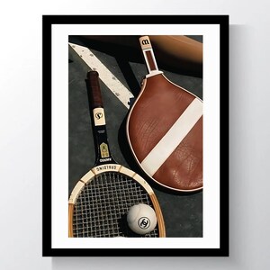 May include: A framed image featuring vintage tennis equipment. The image includes a wooden tennis racket, a brown and white paddle, and a tennis ball with a logo. The equipment is arranged on a dark surface with white lines.
