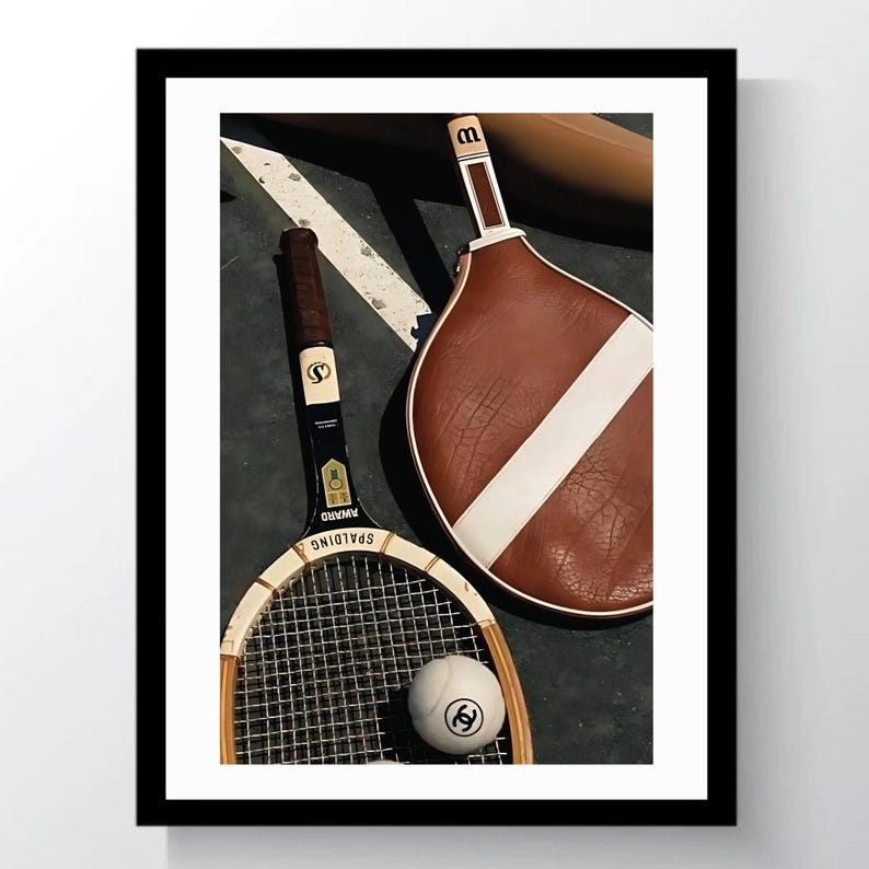 May include: A framed image featuring vintage tennis equipment. The image includes a wooden tennis racket, a brown and white paddle, and a tennis ball with a logo. The equipment is arranged on a dark surface with white lines.