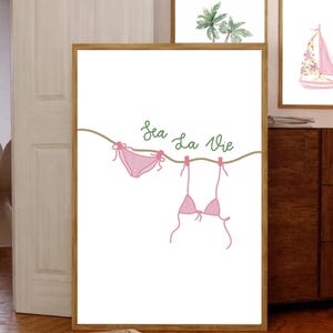 Könnte beinhalten: Ein gerahmter Druck mit einer minimalistischen Illustration eines rosa Bikinis, der an einer Wäscheleine hängt. Die Worte "Sea La Vie" sind in grüner Schreibschrift über dem Bikini geschrieben. Der Hintergrund ist weiß.