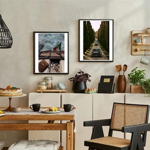 May include: Two framed art prints adorn a neutral wall. One depicts a boat with an Italian flag, the other a road lined with trees and cars. The room includes a wooden table, black mugs, and a woven chair.