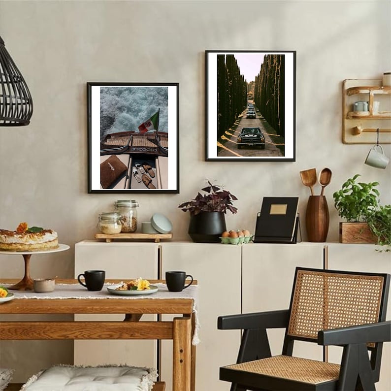May include: Two framed art prints adorn a neutral wall. One depicts a boat with an Italian flag, the other a road lined with trees and cars. The room includes a wooden table, black mugs, and a woven chair.