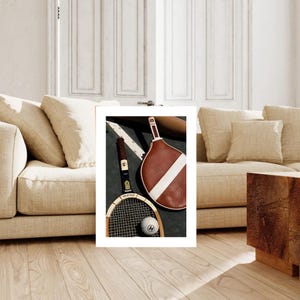May include: A still-life image featuring two vintage tennis rackets and a tennis ball. One racket is brown with a white stripe, the other is black and gold. The tennis ball is white with a black logo. The rackets and ball are on a green surface.