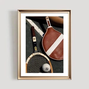 May include: A framed print featuring vintage tennis equipment. The image includes a wooden tennis racket, a brown and white paddle, and a tennis ball with a logo. The background is a dark green surface with white lines.