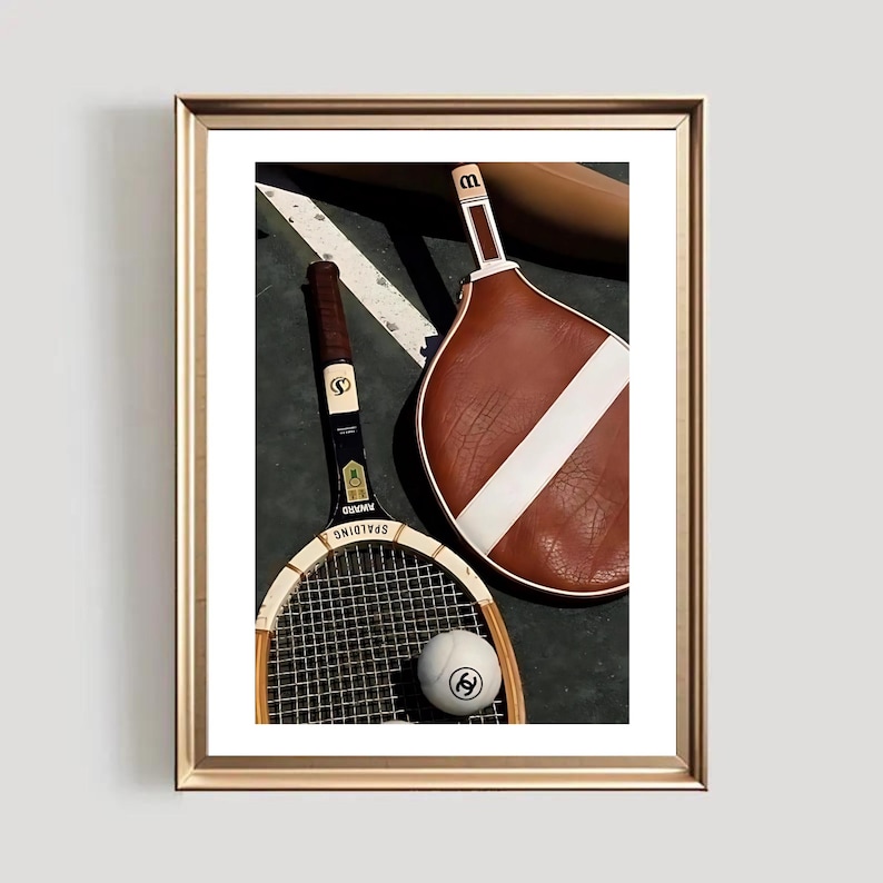 May include: A framed print featuring vintage tennis equipment. The image includes a wooden tennis racket, a brown and white paddle, and a tennis ball with a logo. The background is a dark green surface with white lines.