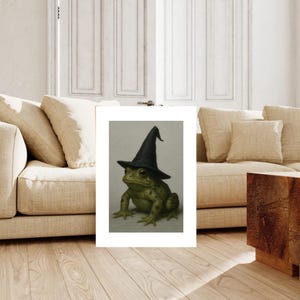 May include: A framed print depicting a green frog adorned with a black witch's hat. The frog, with detailed skin, is centered against a light grey backdrop. The artwork is presented in a white frame, ideal for home decoration.