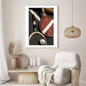 May include: A framed print featuring vintage tennis equipment. The image includes a wooden tennis racket, a brown leather racquet cover with a white stripe, and a tennis ball. The racket has the word "Spalding" on it.