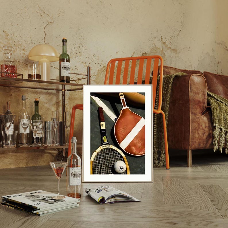 May include: A framed image featuring a tennis racket, a paddleball racket, and a tennis ball on a court. A bottle of wine and a bottle of champagne are also visible. The image is set against a backdrop of a bar cart, a chair, and a leather couch.