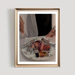 May include: A framed image showing a selection of cosmetics on a decorative plate. The plate contains lipsticks, compacts, and perfume bottles. The frame is gold, and the image is set against a white background.