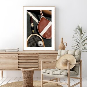 May include: A framed print featuring vintage tennis equipment. Two rackets, one brown and one with a wooden frame, rest beside a tennis ball. The rackets are positioned on a dark surface with a white stripe, suggesting a court setting.