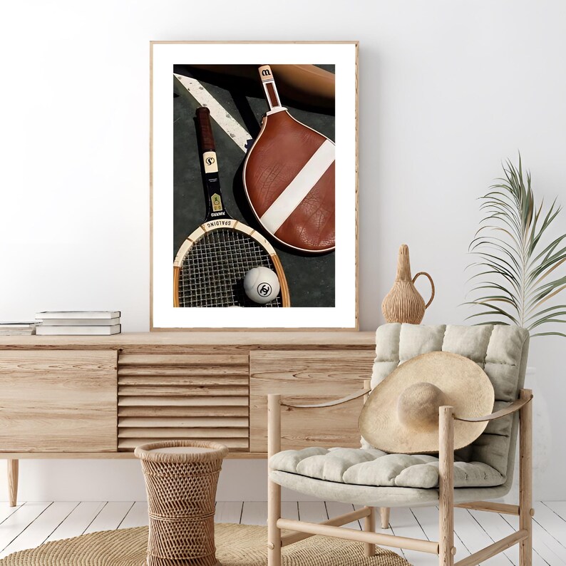 May include: A framed print featuring vintage tennis equipment. Two rackets, one brown and one with a wooden frame, rest beside a tennis ball. The rackets are positioned on a dark surface with a white stripe, suggesting a court setting.