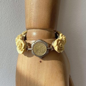 May include: A vintage bracelet featuring cream-colored rose accents and a central medallion. The medallion has a gold-toned center with a detailed design, framed by a silver-toned border. The bracelet is displayed on a wooden mannequin hand.