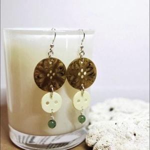 May include: Handmade earrings featuring a unique button design. Each earring has a large, brown, patterned button at the top, followed by a smaller, off-white button, and a small green bead. The earrings hang from silver-colored hooks.