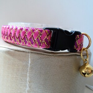 May include: Pink and gold cat collar with a black buckle and a gold bell. The collar is made of a woven ribbon with a checkered pattern.
