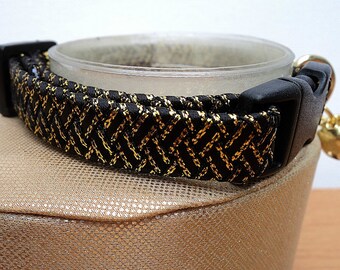 May include: A black and gold cat collar with a woven pattern. The collar has a black plastic buckle and a small gold bell attached. The collar is resting on a gold-colored surface.