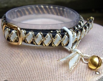 May include: A pet collar featuring a braided pattern of black and white ribbon accented with metallic gold thread. The collar includes a polished gold-tone metal buckle, a white plastic adjustment slider, and a small gold-tone jingle bell charm.