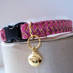 May include: Pink and gold cat collar with a bell. The collar is made of a soft, woven fabric with a gold trim. The bell is gold and has a round shape.