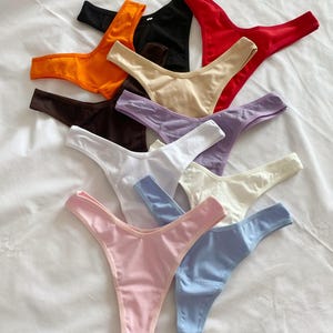 Puede incluir: Una colección de tangas de colores variados, incluyendo naranja, negro, rojo, beige, morado, blanco, rosa y azul. Las prendas están dispuestas sobre una superficie blanca, mostrando sus diferentes colores y estilos.