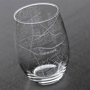 May include: A clear glass wine tumbler with an etched design of the city of Newark, New Jersey. The word "newark." is etched in the center of the glass.
