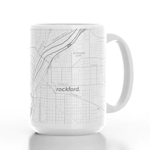 K&ouml;nnte beinhalten: Eine wei&szlig;e Keramiktasse mit einer schwarz-wei&szlig;en Karte von Rockford, Illinois, die darauf gedruckt ist. Die Karte zeigt die Stra&szlig;en der Stadt und den Rock River. Der Text "rockford." ist in Schwarz unter der Karte gedruckt.