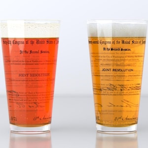 May include: Two clear glass pint glasses with a printed design of the text of the 18th Amendment to the United States Constitution. The glass on the left is filled with a red liquid and the glass on the right is filled with a yellow liquid.