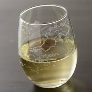 May include: A clear glass wine glass with an etched design of the Virgin Islands. The glass is filled with a light yellow liquid and the text "st john." is etched on the bottom of the glass.
