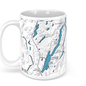 May include: White ceramic mug with a map of Lake George, New York printed on it. The map shows the lake and surrounding area, including the towns of Bolton, Lake George, and Ticonderoga. The map is in black and white with blue water features.