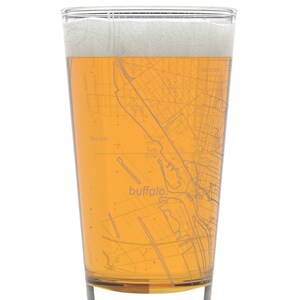 May include: A clear glass pint glass with a map of Buffalo, New York etched on the side. The glass is filled with a light amber beer with a white head.