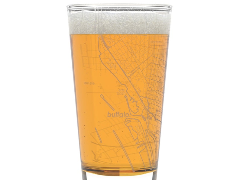 May include: A clear glass pint glass with a map of Buffalo, New York etched on the side. The glass is filled with a light amber beer with a white head.
