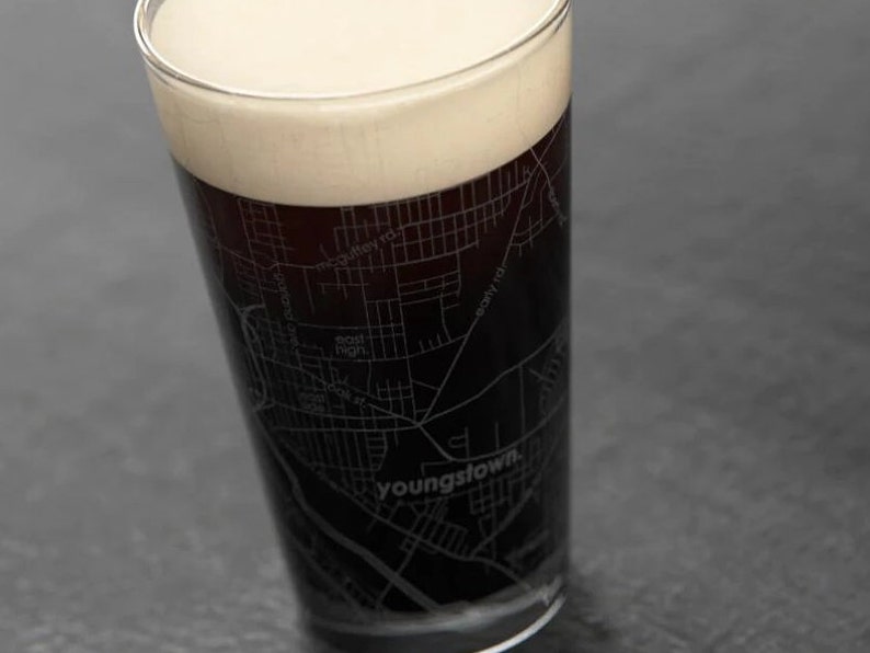 May include: A pint glass with a black and white map of Youngstown, Ohio, etched into the side. The glass is filled with a dark beer with a thick head of foam.