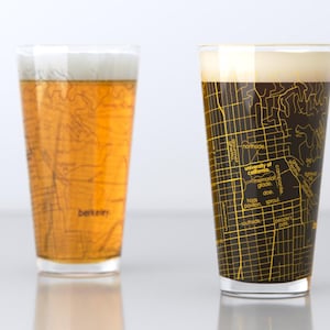 May include: Two clear glass pint glasses with a map of Berkeley, California printed on the bottom. The glass on the left is filled with a light-colored beer, while the glass on the right is filled with a dark-colored beer.