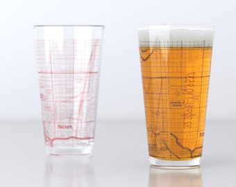 May include: Two clear glass pint glasses with a grid pattern printed on the side. The glass on the left is empty, while the glass on the right is filled with a light amber liquid. The grid pattern on the glass on the right is filled with a map of Tucson, Arizona, with the text "University of Arizona" printed on the map.