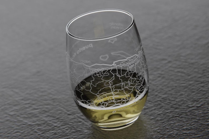 May include: A clear glass wine glass with an etched design of the city of San Juan, Puerto Rico. The glass is filled with a light yellow liquid.
