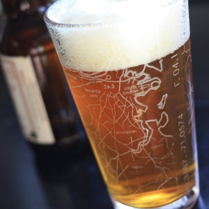 May include: A pint glass filled with beer and foam, featuring an etched map design. The glass is partially filled with a light amber liquid. A brown bottle is in the background.