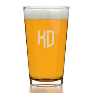 May include: A clear glass pint glass filled with a light yellow beer. The glass has a white monogram of the letters "KD" in a shield shape.
